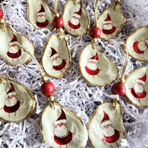 May include: A collection of 12 handmade Christmas ornaments featuring Santa Claus painted on oyster shells. Each ornament is adorned with a red bead and gold trim.