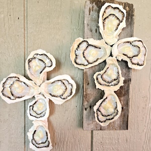 May include: Two oyster shell crosses, each made of five oyster shells, arranged in a cross shape. The shells are white with black edges and are mounted on a piece of weathered wood.