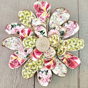 May include: A decorative flower made from painted oyster shells. The shells are adorned with floral patterns in pink, red, green, and white. The center of the flower is a coil of natural twine. The shells are arranged on a wooden surface.