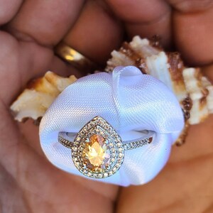 May include: A pear-shaped engagement ring with a large, golden-yellow center stone surrounded by smaller, clear stones. The ring is set in a silver band and is displayed on a white satin ribbon.