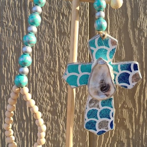 May include: A wooden cross with a blue and silver seashell design and an oyster shell in the center. The cross is hanging from a wooden dowel with a turquoise and silver beaded garland.