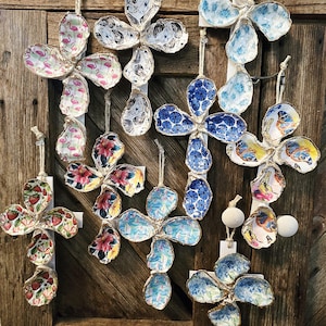 May include: A collection of decorative oyster shell ornaments in a variety of designs. The ornaments are shaped like flowers and feature painted patterns, including flamingos, floral prints, and blue and white designs. They hang from twine.
