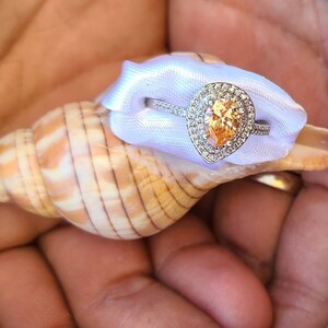 May include: A pear-shaped engagement ring with a yellow center stone and a white gold band, displayed on a seashell and a white satin ribbon.