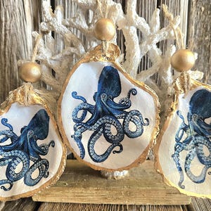May include: Three oyster shell ornaments with a gold rim and a blue octopus design. Each shell is suspended by a gold string and bead. The background features a white coral and weathered wood.