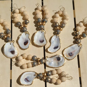 May include: Seven oyster shell ornaments with gold trim and a wooden bead garland in gray and gold. The ornaments are hanging from a wooden surface.