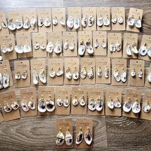 May include: A collection of 40 pairs of oyster shell earrings with gold accents. The earrings are displayed on brown card stock with the text "Handmade with Love" printed on each card.