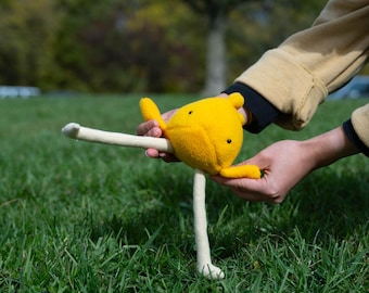 May include: A handmade yellow plush toy with black dot eyes and a beak, held in a person's hands against a backdrop of green grass and a blurred tree. The toy has a long, cream-colored leg and a small tail.