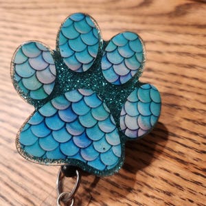 May include: A teal blue paw print with a mermaid scale design and glitter accents. The paw print is attached to a clear plastic retractable badge holder.