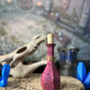 May include: A small, dark pink glass bottle with a cork stopper. The bottle has a textured surface and is tied with twine. The bottle is sitting on a wooden surface.