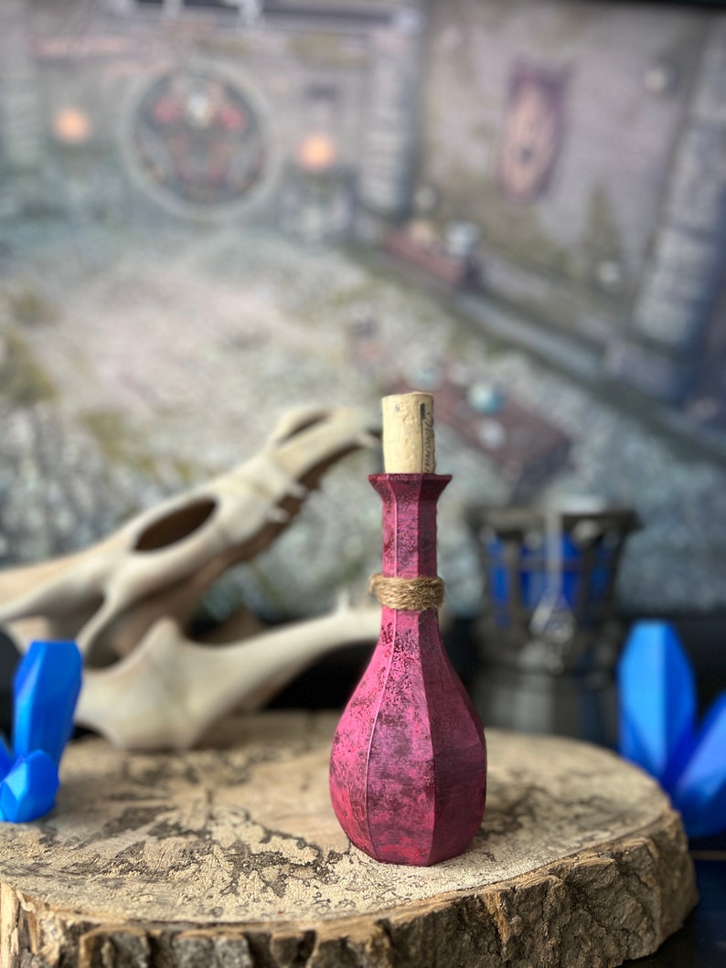 May include: A small, dark pink glass bottle with a cork stopper. The bottle has a textured surface and is tied with twine. The bottle is sitting on a wooden surface.