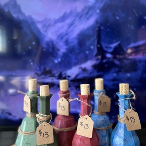 May include: Six small glass bottles with cork stoppers and brown paper tags with the price $13 written on them. The bottles are filled with different colored liquids and have a textured, faceted design. The bottles are arranged in a row on a wooden surface.
