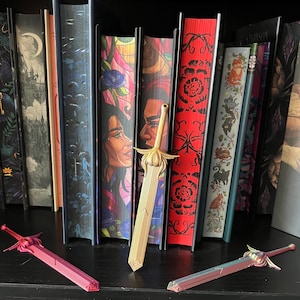 Puede incluir: Tres espadas decorativas doradas con diseños intrincados, descansando sobre un estante negro frente a una estantería llena de libros coloridos.