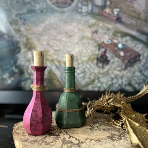 May include: Two small glass bottles with cork stoppers, one is pink and the other is green, sitting on a wooden surface with a gold dragon figurine.