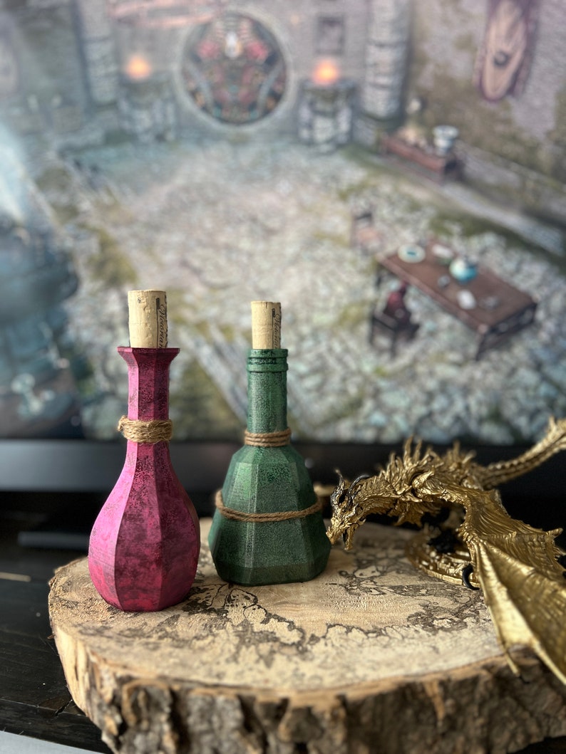 May include: Two small glass bottles with cork stoppers, one is pink and the other is green, sitting on a wooden surface with a gold dragon figurine.