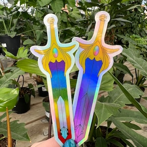 Puede incluir: Dos pegatinas holográficas iridiscentes de una espada con mango dorado y hoja azul, púrpura y arcoíris.