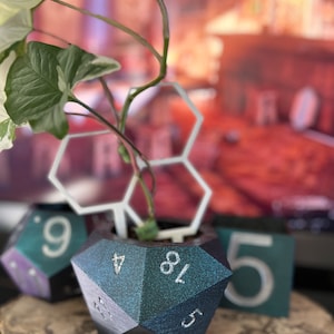 Puede incluir: Un macetero de dados d20 de color azul verdoso y negro iridiscente con números blancos. El macetero está lleno de una pequeña planta verde y un soporte hexagonal blanco. El macetero está sobre una superficie de madera.