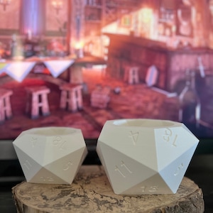 May include: Two white, geometric dice-shaped planters with numbers on the sides. The planters are on a wooden slice. The background is blurred, showing a room with a bar and stools.