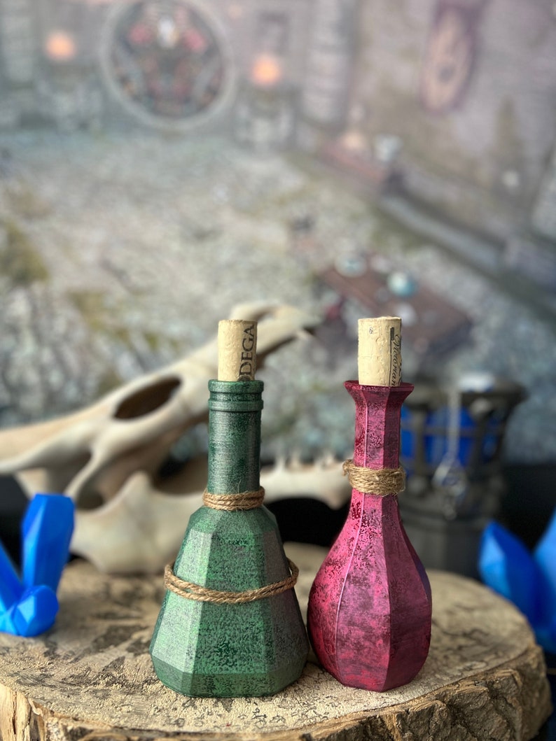 May include: Two small glass bottles with cork stoppers. The bottle on the left is green and the bottle on the right is pink. Both bottles have a rope tied around the neck.