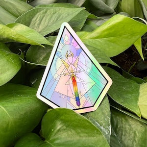 Op de afbeelding: Een holografische sticker met een geometrisch ontwerp met een regenboogzwaard en een figuur met vleugels. De sticker heeft de vorm van een driehoek.