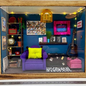 May include: A miniature room diorama featuring a purple armchair, a yellow cushion, a guitar, a record player, and a neon sign reading "Happy Hour". The room is decorated with a blue wall, a shelf with books and plants, and a string of fairy lights.