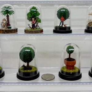 May include: Eight miniature glass domes containing various scenes. The domes are arranged in two rows on a clear shelf. The scenes include a palm tree, a woman with a bicycle, a man standing, a tree with a cow, a tree with a cat, a tree with a potted plant, a tree with a bird, and a tree with a mushroom.