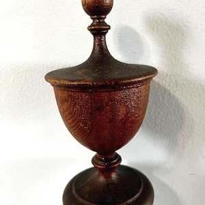 May include: A dark brown wooden urn with a textured grain finish, featuring a rounded base, a tapered stem, a wide bowl-shaped body, and a matching lid topped with a spherical finial. The object displays a deep, rich wood tone throughout.
