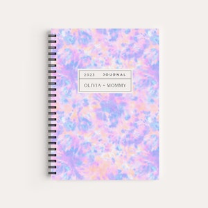 May include: A spiral bound journal with a tie-dye cover in shades of pink, blue, and white. The cover has the text "2023 JOURNAL" and "OLIVIA + MOMMY" printed on it.