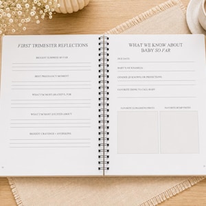 May include: An open spiral-bound pregnancy journal with white pages featuring black text. The left page is titled FIRST TRIMESTER REFLECTIONS with prompts for surprises, moments, gratitude, excitement, and cravings. The right page is titled WHAT WE KNOW ABOUT BABY SO FAR with prompts for due date, nickname, gender, and two blank squares for photos.