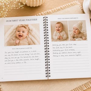 May include: A spiral-bound baby memory book open to two pages titled Our First Year Together and What I'm Looking Forward To. The pages feature printed photo frames with images of a smiling baby, lined sections for handwritten notes, and text.