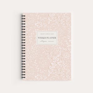 May include: A spiral-bound weekly planner with a light pink cover featuring a white embossed floral pattern. A central rectangular label displays the text SMART SIMPLE MOM, WEEKLY PLANNER, and allyson marie in black lettering on a white background.