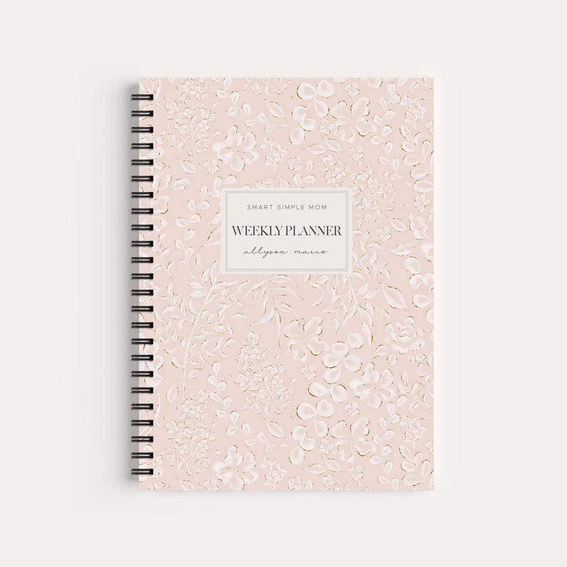May include: A spiral-bound weekly planner with a light pink cover featuring a white embossed floral pattern. A central rectangular label displays the text SMART SIMPLE MOM, WEEKLY PLANNER, and allyson marie in black lettering on a white background.