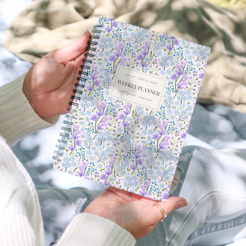 May include: A spiral-bound weekly planner featuring a floral pattern of light blue and purple flowers on a white background. A central rectangular label displays the text "SMART SIMPLE MOM", "WEEKLY PLANNER", and the name "allyson marie" in black.