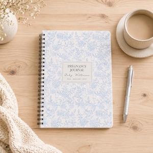 May include: A spiral-bound pregnancy journal with a light blue and white floral pattern on the cover. The center label features the text PREGNANCY JOURNAL, Baby Williams, and DUE JANUARY 2025. The journal is positioned next to a silver pen.