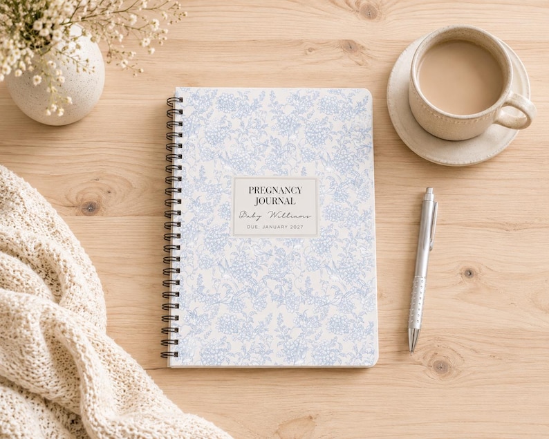 May include: A spiral-bound pregnancy journal with a light blue and white floral pattern on the cover. The center label features the text PREGNANCY JOURNAL, Baby Williams, and DUE JANUARY 2025. The journal is positioned next to a silver pen.