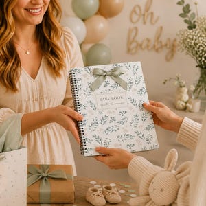 May include: A spiral-bound baby book featuring a white cover with a green botanical leaf pattern, a sage green ribbon bow at the top, and a rectangular label with the text BABY BOOK printed in black ink on the front cover.