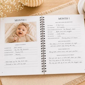 May include: A spiral-bound baby memory book open to a page titled Month 1. The page features a photograph of a smiling infant wearing a bow headband, alongside handwritten entries detailing weight as 8 lbs 2 oz, sleep habits, and milestones.