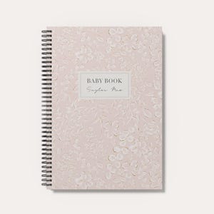 May include: A spiral-bound baby book with a light pink cover featuring a subtle, tonal floral and leaf pattern. A rectangular white label in the center displays the text BABY BOOK in a bold font and the name Saylor Mae in a script font.