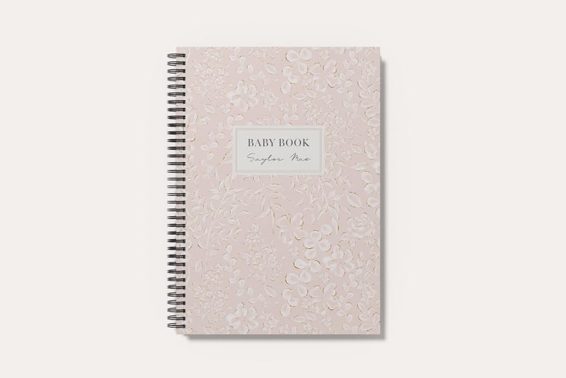 May include: A spiral-bound baby book with a light pink cover featuring a subtle, tonal floral and leaf pattern. A rectangular white label in the center displays the text BABY BOOK in a bold font and the name Saylor Mae in a script font.