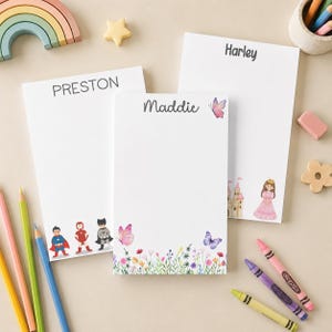 May include: Three rectangular white paper notepads with personalized names printed at the top. One notepad features superhero illustrations, one features a floral and butterfly design with the name Maddie, and one features a princess and castle.