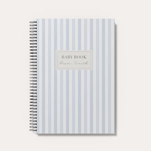 May include: A spiral-bound notebook featuring a light blue and white vertical striped pattern. Centered on the cover is a rectangular label with a light gray border containing the text BABY BOOK and the name Liam Smith in a script font.