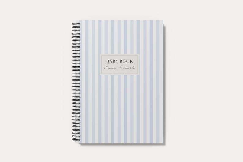 May include: A spiral-bound notebook featuring a light blue and white vertical striped pattern. Centered on the cover is a rectangular label with a light gray border containing the text BABY BOOK and the name Liam Smith in a script font.
