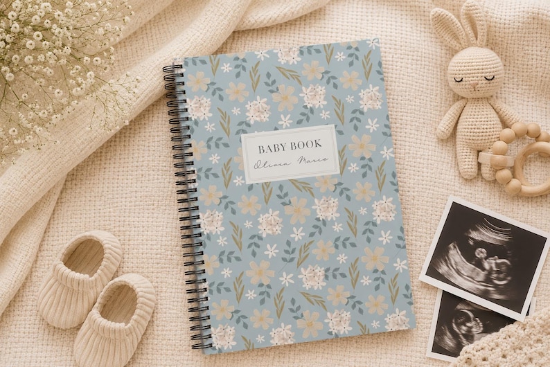 May include: A spiral-bound baby book with a light blue cover featuring a floral pattern of white and beige flowers with green leaves. A white rectangular label on the front displays the text BABY BOOK and the name Olivia Marco in black font.