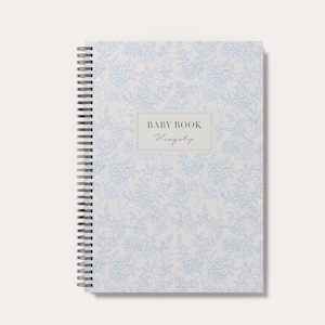 May include: A spiral-bound baby book with a light gray cover featuring a repeating blue floral pattern. A central rectangular label displays the text BABY BOOK and the name Kingsly in a script font. The notebook has a black metal coil spine.