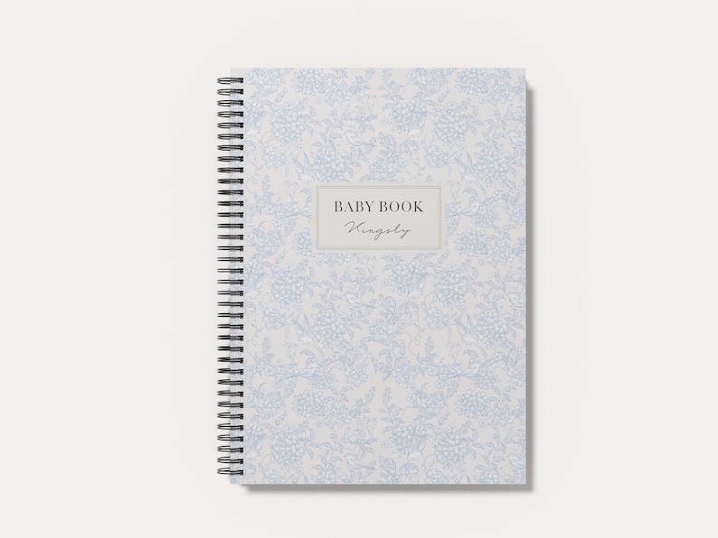 May include: A spiral-bound baby book with a light gray cover featuring a repeating blue floral pattern. A central rectangular label displays the text BABY BOOK and the name Kingsly in a script font. The notebook has a black metal coil spine.