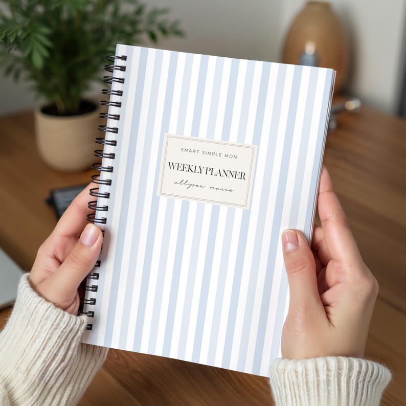 May include: A spiral-bound weekly planner featuring a light blue and white vertical striped pattern. The cover includes a central rectangular label with the text SMART SIMPLE MOM, WEEKLY PLANNER, and allyson marie printed in a dark font.