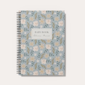 May include: A spiral-bound notebook with a light blue cover featuring a repeating pattern of small white and beige flowers with green leaves. A central rectangular label displays the text BABY BOOK and a script name in black ink.
