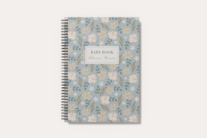 May include: A spiral-bound notebook with a light blue cover featuring a repeating pattern of small white and beige flowers with green leaves. A central rectangular label displays the text BABY BOOK and a script name in black ink.