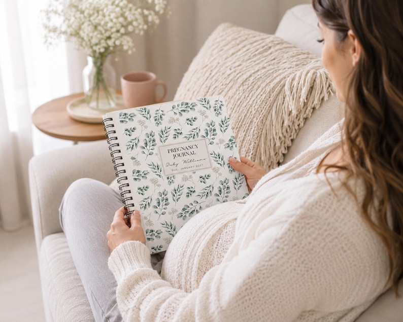 May include: A spiral-bound pregnancy journal with a white cover featuring a green botanical leaf pattern. The center label displays the text PREGNANCY JOURNAL, Baby Williams, and DUE JANUARY 2027 in black ink. The journal has a black wire binding.