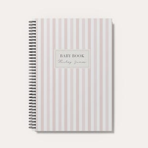 May include: A spiral-bound notebook featuring a vertical light pink and white striped pattern. The center of the cover displays a rectangular label with the text BABY BOOK and the name Harley James printed in a dark gray serif and script font.