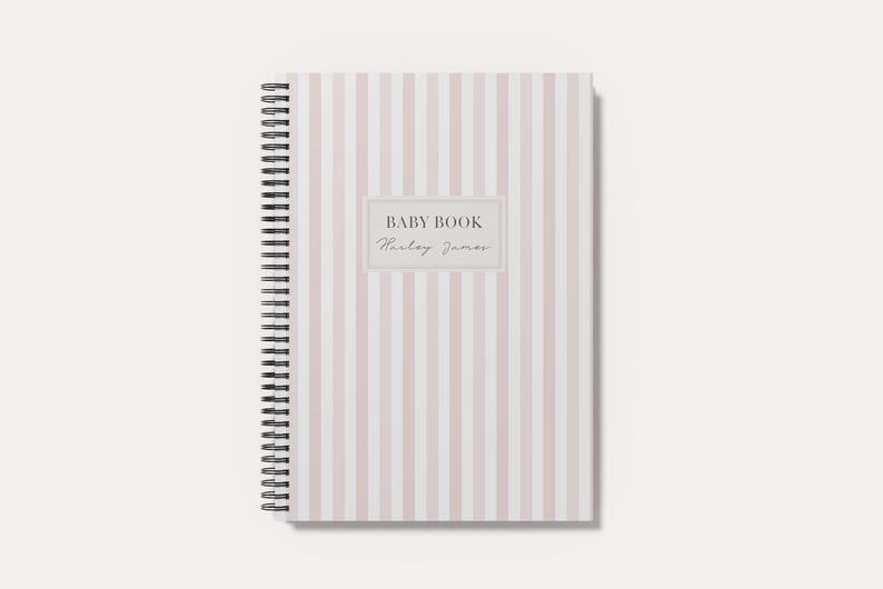 May include: A spiral-bound notebook featuring a vertical light pink and white striped pattern. The center of the cover displays a rectangular label with the text BABY BOOK and the name Harley James printed in a dark gray serif and script font.